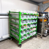 A green metal trolley racking unit with multiple shelves, holding white plastic bins, is placed in an industrial setting.